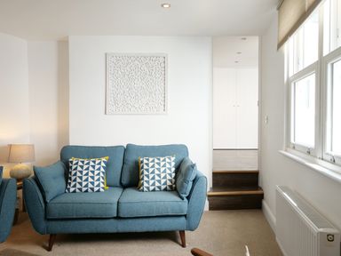 A living room with a blue sofa and patterned cushions beside a lamp and window at Waves End in St Ives