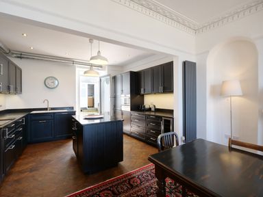 A kitchen with dark cabinets a central island a dining table a floor lamp and a wall clock at Waves End in St Ives