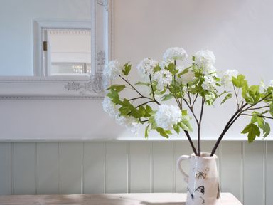 A vase with white flowers and green leaves on a wooden table with a decorative white framed mirror on the wall