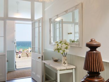 An entryway with a wooden table holding a vase of flowers and a bowl a large mirror on the wall and an open door showing people sitting by the seaside at Waves End in St Ives