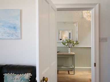 A room with a wooden table holding a white vase with flowers a mirror on the wall and a partially open door leading to a room with a black couch and a blue pillow at Waves End St Ives