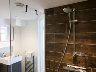 A bathroom shower area with a handheld showerhead and dark wood-effect tiles at Waves End in St Ives