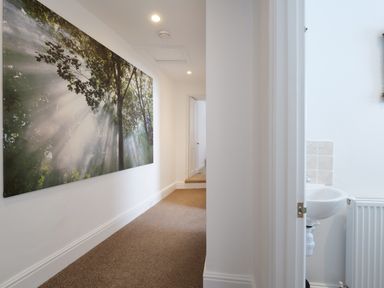 A hallway with a large wall art of sunlight through trees and a partially visible bathroom with a sink and radiator at Waves End in St Ives