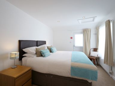 A bedroom with a bed blue pillows a wooden bedside table with a lamp and a chair by the window at Waves End in St Ives
