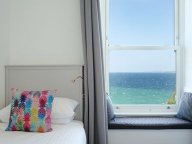 A bedroom with a bed and colorful pillow beside a window seat overlooking the ocean at Waves End in St Ives