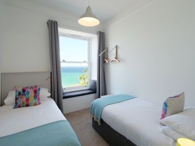 A bedroom with two single beds pillows with colorful covers window seat with cushions and a view of the sea at Waves End in St Ives