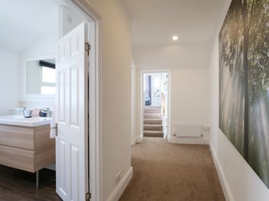 A hallway with carpet leading to a bedroom and a bathroom with a wooden vanity unit and mirror