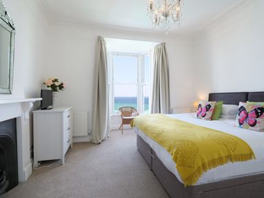 A bedroom with a double bed yellow throw pillows with butterflies a cabinet with a TV and flowers and a window with ocean view at Waves End in St Ives