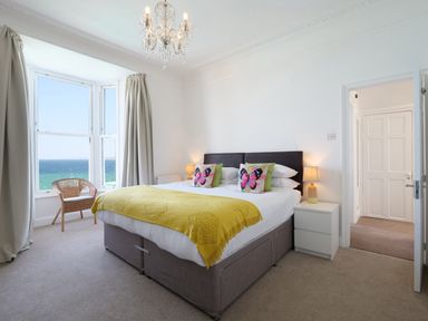A bedroom with a large bed yellow throw pillows a chair by the window and a view of the sea at Waves End in St Ives