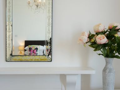 A mirror above a white table reflecting a bed with a butterfly cushion and a lamp next to a vase with flowers at Waves End in St Ives