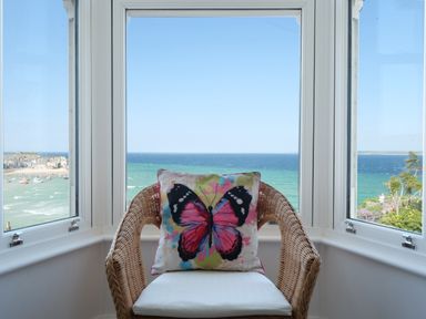 A wicker chair with a butterfly cushion in front of a bay window overlooking the ocean at Waves End in St Ives