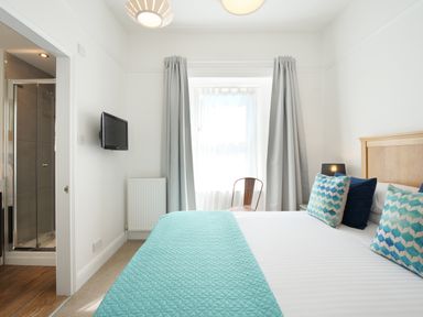 A bedroom with a bed with blue and white bedding a television on the wall and a chair by the window at Waves End in St Ives