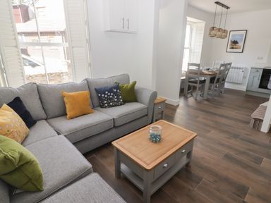A living room with a grey sofa with cushions and a wooden coffee table next to a dining area with a wooden table and chairs at Gwynfryn in Pen-Clawdd