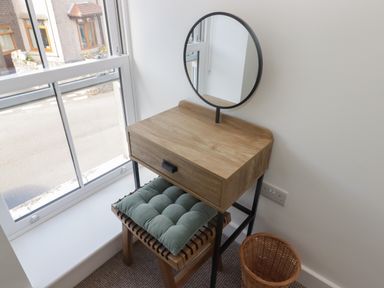 A small wooden vanity table with a round mirror and a cushioned stool next to a window and a wicker basket on a carpeted floor at Gwynfryn in Pen-Clawdd