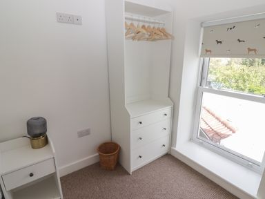 A corner of a room with a white dresser with hangers a window with a dog patterned blind and a small nightstand with a lamp at Gwynfryn in Pen-Clawdd