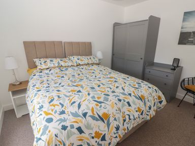 A bedroom with a double bed with floral bedding a gray wardrobe gray chest of drawers and a chair at Gwynfryn in Pen-Clawdd