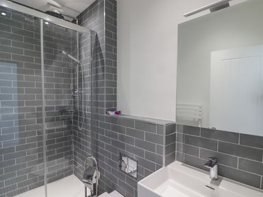 A bathroom with a glass shower enclosure gray tiled walls a white sink and a large mirror at Gwynfryn in Pen-Clawdd
