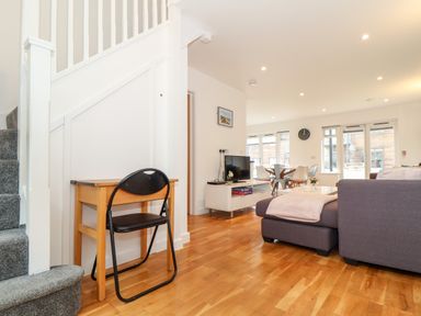 A living area with a small desk and chair under the stairs a gray couch a TV on a white stand and a dining table with chairs at Garras in Porthtowan