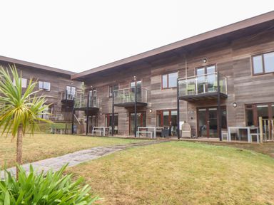 A grassy courtyard area with a stone pathway in front of two story buildings with wooden exteriors and balconies at Garras in Porthtowan