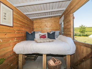 A wooden cabin interior with a bed elevated on wooden legs with pillows and a basket of firewood underneath at The Snug in Dulverton