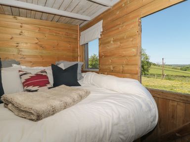 A bed with pillows and a blanket inside a wooden room with a window showing a green field at The Snug in Dulverton