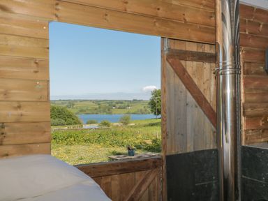 A wooden room with a bed and an open door showing a view of a grassy field and a body of water at The Snug in Dulverton