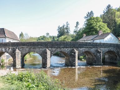A stone arch bridge over a shallow river with houses and trees in the background at The Snug in Dulverton