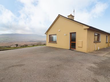 A single-story house with yellow walls and several windows on a paved area with hills in the background at Gortnamona in Coolnaharragill Lower near Glenbeigh County Kerry