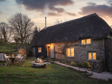 A stone cottage with a thatched roof and lit windows in a garden with outdoor seating at Butterwell Cottage at Collihole in Chagford