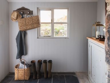 An entryway with a window a wooden coat rack with hats and bags rubber boots on the floor a basket with blankets and a cabinet with a lantern and candle at Butterwell Cottage at Collihole in Chagford