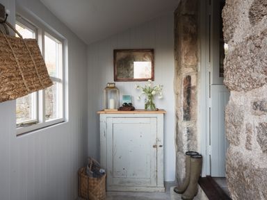 A hallway with a white cabinet topped with a lantern, flowers, and binoculars next to stone walls and boots by the door at Butterwell Cottage at Collihole in Chagford