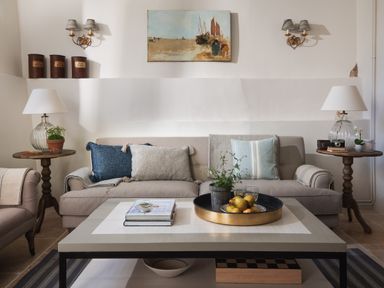 A living room with a beige sofa blue and gray cushions wooden side tables with lamps a coffee table with a tray of fruit and books at Butterwell Cottage at Collihole in Chagford