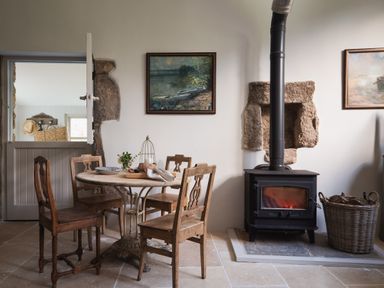 A dining area with a wooden table and chairs near a wood burning stove with a basket of firewood at Butterwell Cottage at Collihole in Chagford