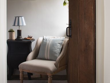 A beige armchair with a striped cushion next to a black bedside table with a lamp plant and glasses at Butterwell Cottage at Collihole in Chagford