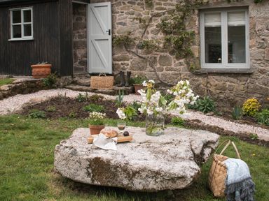A stone table with flowers in a vase and food on it outside a stone house with a garden and a wicker basket on grass at Butterwell Cottage at Collihole in Chagford