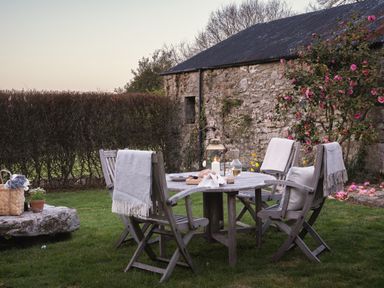 An outdoor dining area with wooden chairs and a round table set on grass near a stone building with pink flowers at Butterwell Cottage at Collihole in Chagford