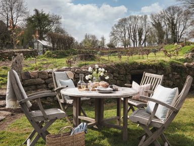 An outdoor wooden table with four chairs on grass with bread flowers and glasses near a stone wall and trees at Butterwell Cottage at Collihole in Chagford