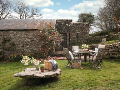 An outdoor garden area with wooden chairs around a table beside a stone wall and a large flat stone with flowers and a basket on it at Butterwell Cottage at Collihole in Chagford