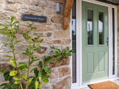 A green front door with glass panels next to a stone wall with a plant and a sign that reads Chynoweth House at Chynoweth House in Cubert