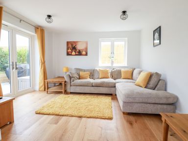 A living room with a gray sectional sofa with yellow cushions a yellow rug wooden tables a lamp and framed pictures at Chynoweth House in Cubert