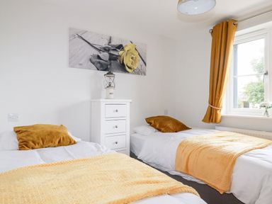 A bedroom with two single beds yellow blankets and pillows a white chest of drawers a window with yellow curtains and a flower painting on the wall at Chynoweth House in Cubert