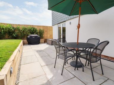 An outdoor patio with a round table and four chairs under a green umbrella next to a grassy area with a wooden fence and a covered barbecue at Chynoweth House in Cubert