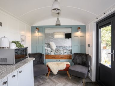 A small living area with two chairs and a bench in front of a built-in bed alcove with a patterned bedspread and a small mounted TV at Shepherd's Stone in Llanfairynghornwy near Llanfaethlu