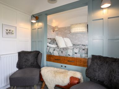 A cozy seating area with two gray chairs and a sheepskin-covered footstool facing a built-in bed with patterned bedding at Shepherds Stone in Llanfairynghornwy near Llanfaethlu