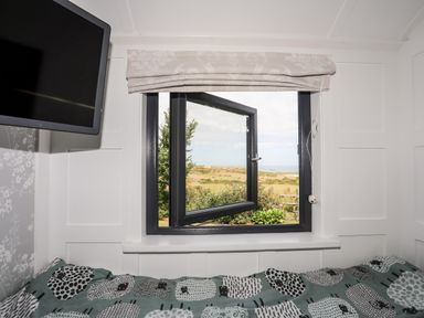 A bedroom with a patterned bedspread a wall-mounted TV and an open window showing an outdoor landscape at Shepherds Stone in Llanfairynghornwy near Llanfaethlu