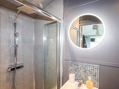 A bathroom with a shower area with glass door a sink with a faucet soap dispenser and a round mirror with light at Shepherd's Stone in Llanfairynghornwy near Llanfaethlu