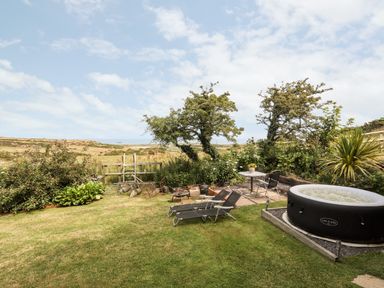 An outdoor garden with two lounge chairs a small table and chairs a hot tub and bushes at Shepherd's Stone in Llanfairynghornwy near Llanfaethlu