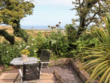 A garden seating area with a table and two chairs surrounded by greenery and a view of the sea at Shepherds Stone in Llanfairynghornwy near Llanfaethlu