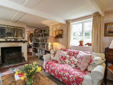 A living room with a sofa covered in a red floral throw pillows a coffee table with a vase of flowers a window a fireplace and a bookshelf at Chalkpit Cottage in Blewbury