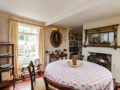 A dining room with a round table covered with a tablecloth and chairs around it and a fireplace with a large mirror above and a bookshelf in the corner at Chalkpit Cottage in Blewbury